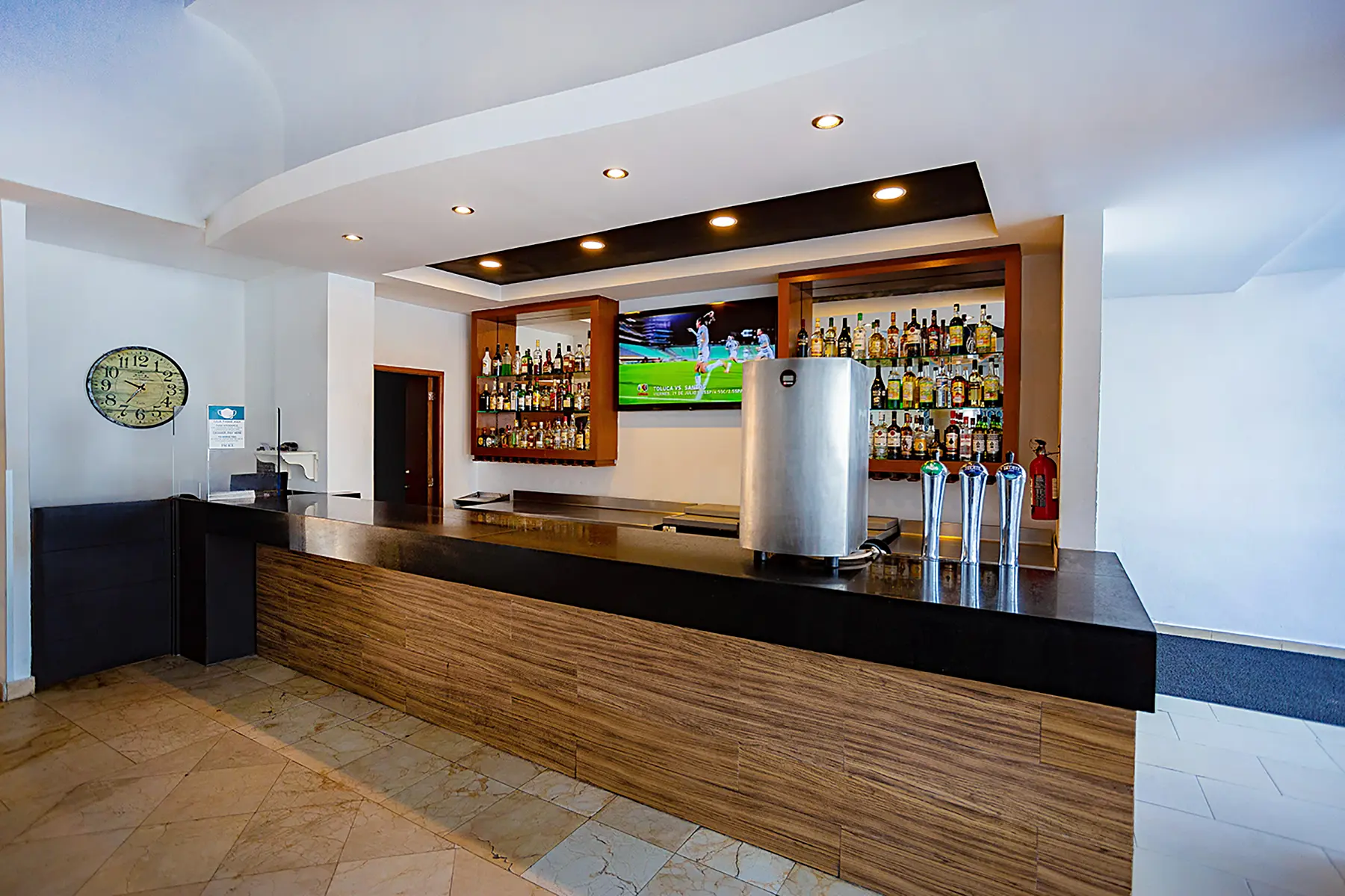 Barra de bebidas de madera, un reloj, botellas de alcohol y una pantalla en Grand Central Sport Bar de hotel Oceano Palace Mazatlán